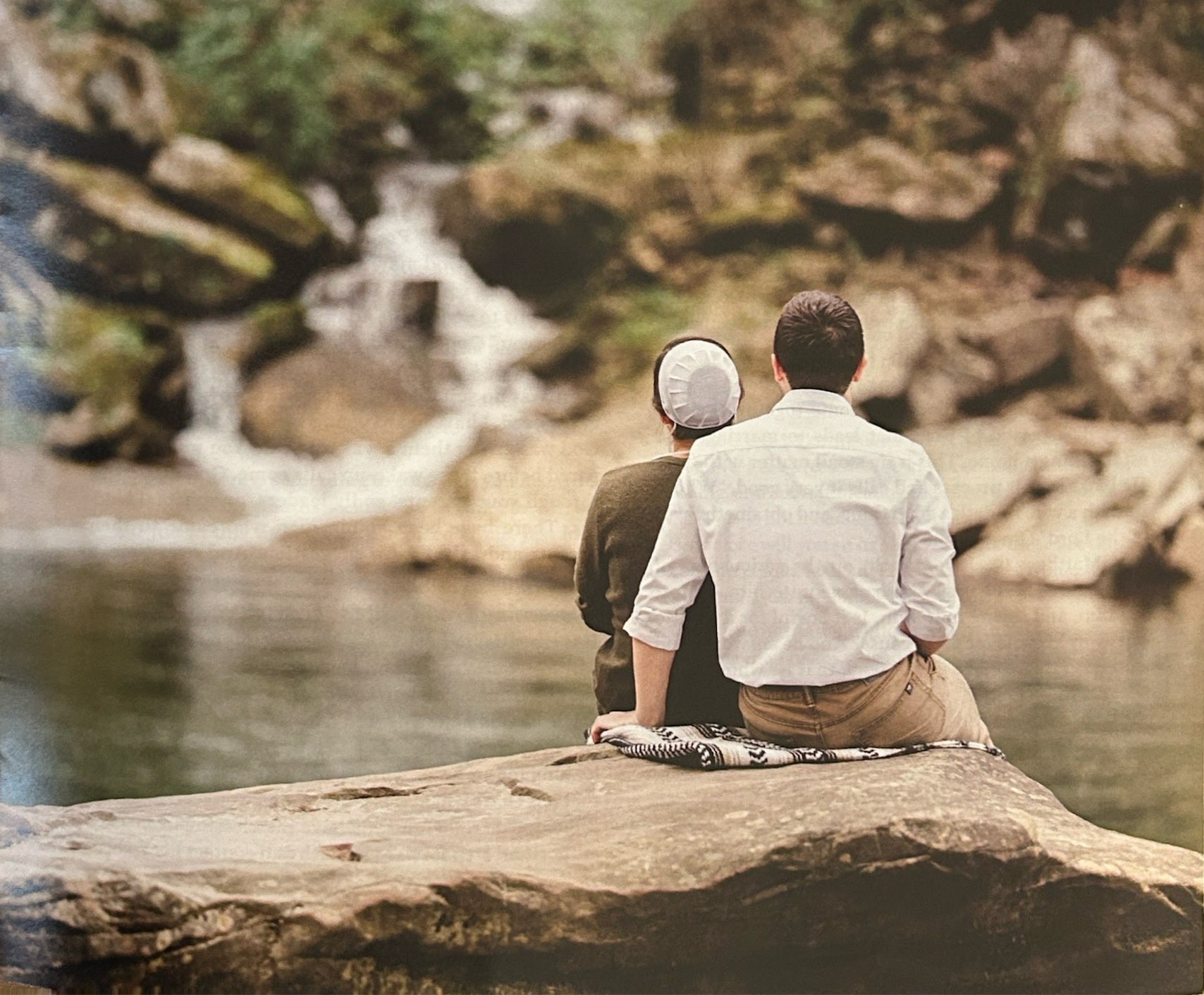 A couple seated together on a rock facing a waterfall.
