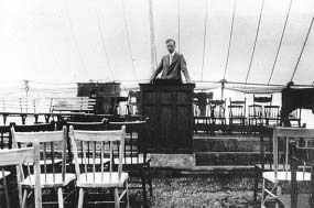A man standing at a pulpit among chairs inside a tent.