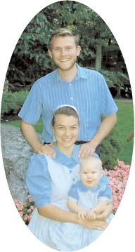 A man standing behind a seated woman holding a baby in a garden.