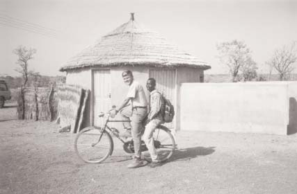 Daniel Kenaston and Matthew with a bicycle beside a round thatched building.