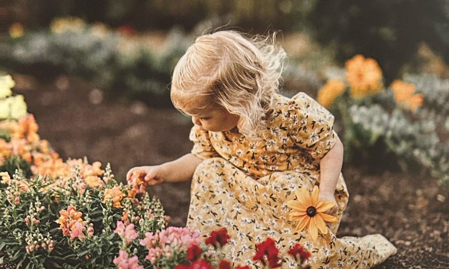 A young child in a yellow floral dress among garden flowers.