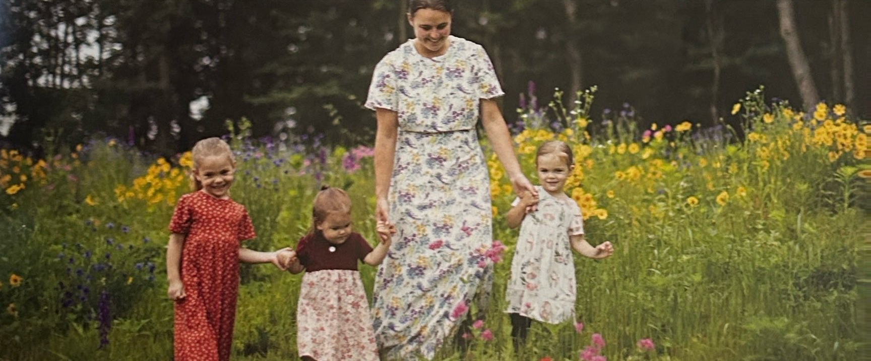 A woman and three young children hold hands among flowers.