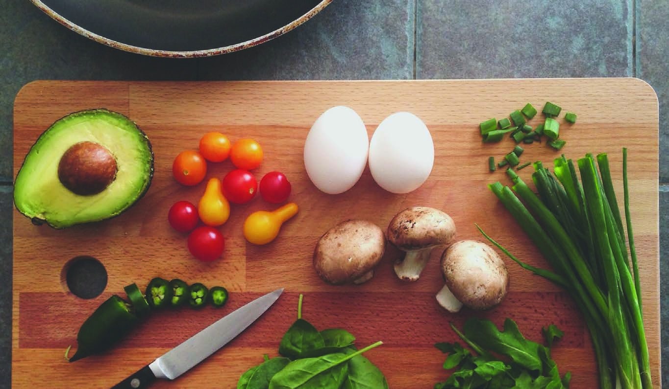 Avocado, eggs, vegetables, mushrooms, and a knife arranged on a cutting board
