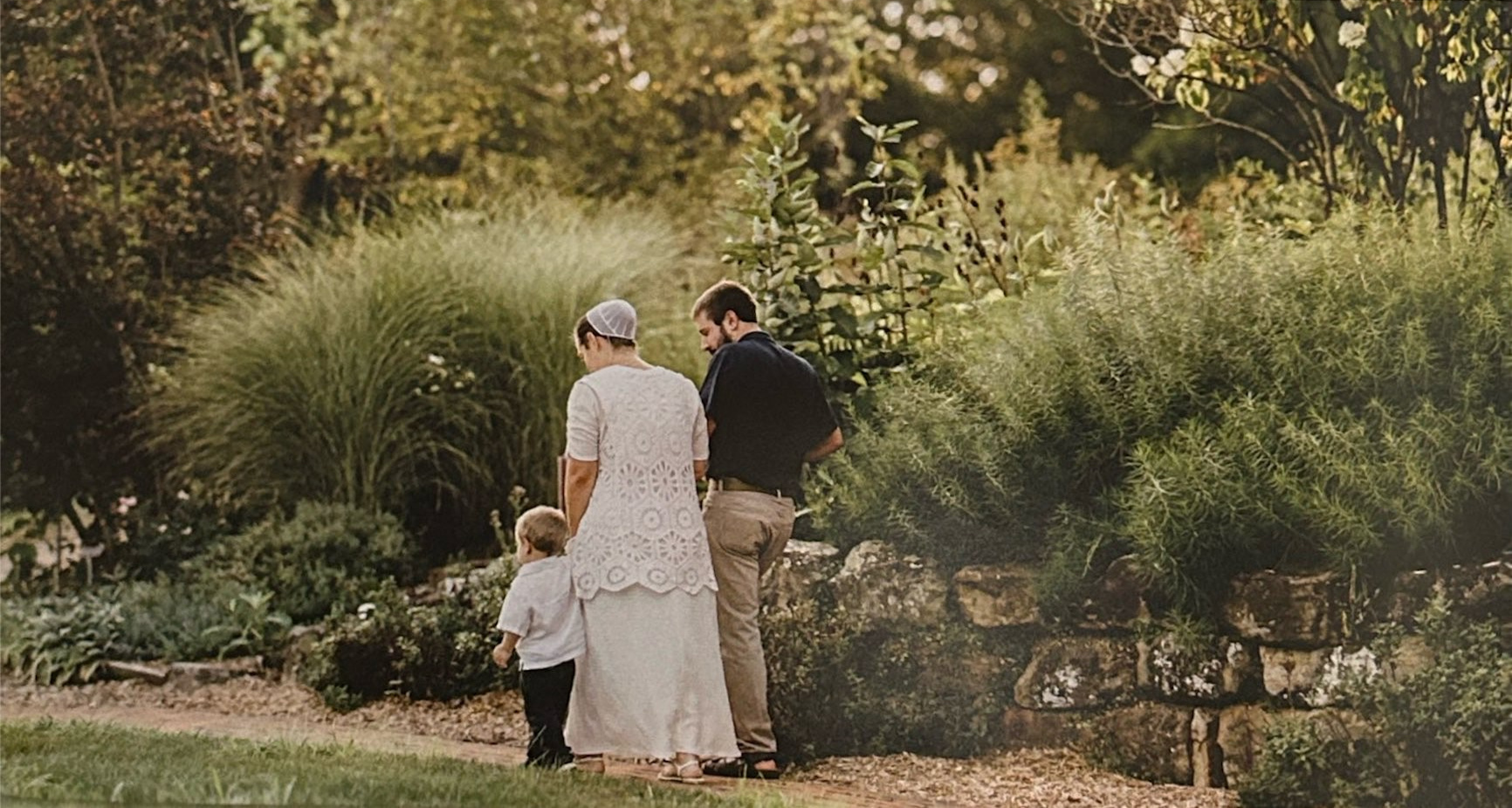 A family walks away along a garden path amid trees and tall grasses.