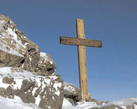 A wooden cross beside snow-covered rocks under a blue sky.