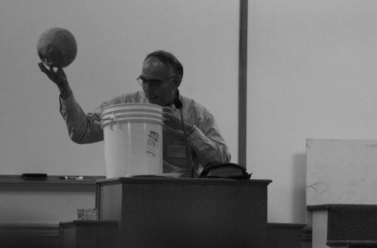 A speaker holding a ball above a bucket at a lectern